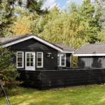 Black wooden house with patio, table, two chairs, and bench. Surrounded by greenery and trees.