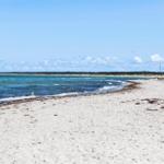 Strand mit grauem Sand und tiefblauem Meer unter klarem Himmel.