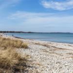 Strand mit Kies und Gras, Blick auf das Meer unter blauem Himmel.