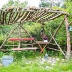 Schwarze Holzhütte mit Garten, Grill und Ofen. Tisch und Stühle auf Steinplatten.