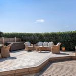 Terrasse mit Sitzgruppe, Tisch und Stühlen. Hintergrund: Hecke und klarer Himmel.