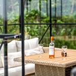 Terrasse mit Glasgarten, Rattanmöbeln und rotem Liegestuhl. Hintergrund: Grasland und Bäume.