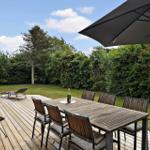 Terrasse mit Holztisch, Stühlen und Sonnenschirm. Grünfläche und Bäume im Hintergrund.
