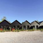 Row of dark wooden cabins with garden tables and clear sky.