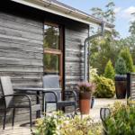 Moderne Holzhütte mit Terrasse, Garten und Garage. Umgeben von Bäumen und Gras.
