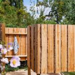 Wooden outdoor shower with towel hooks and green surroundings.