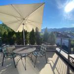 Terrasse mit Tisch, zwei Stühlen und Sonnenschirm. Blick auf grüne Hügel und Häuser.