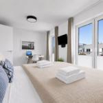Schlafzimmer mit Bett, Schreibtisch und TV. Große Fenster mit Blick auf Balkon.