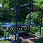 Außenbereich mit Grillunterstand, Tisch und Bank umgeben von Grünflächen.