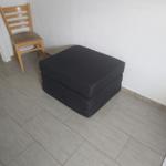 Black ottoman and wooden chair in a room with light walls and tiled floor.